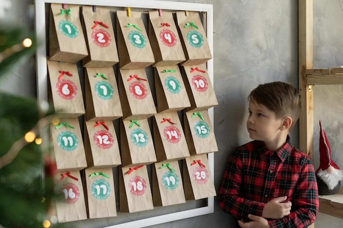 Ręcznie wykonany kalendarz adwentowy wiszący na ścianie, dziecko stojące obok toreb z słodyczami Boże Narodzenie