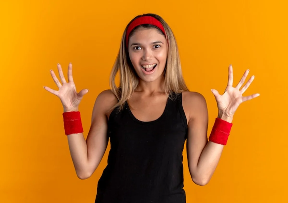 Młoda dziewczyna fitness w czarnej odzieży sportowej i czerwonej opasce, stojąca nad pomarańczową ścianą